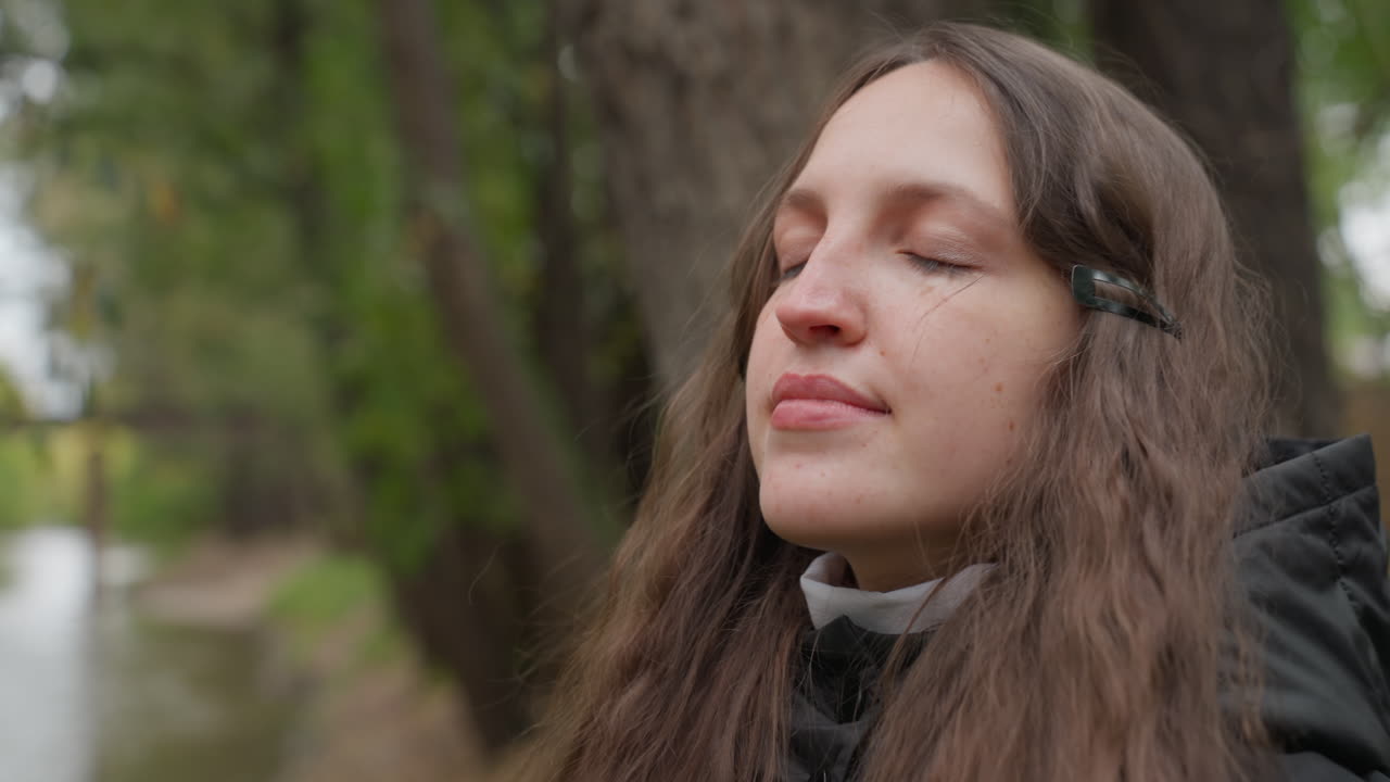 Giovane donna caucasica che percorre un sentiero nel bosco, primi piani che catturano espressioni pensierose vicino al fiume che appare come un'escursionista, una poetessa, una studentessa che riflette vicino al ruscello con foglie autunnali, giacca con cappuccio, lunga