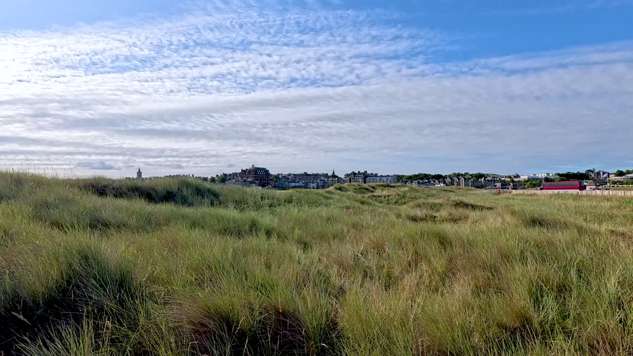 Camera moves toward rustic fence through grassy sand dunes under bright sky, Fife, Scotland