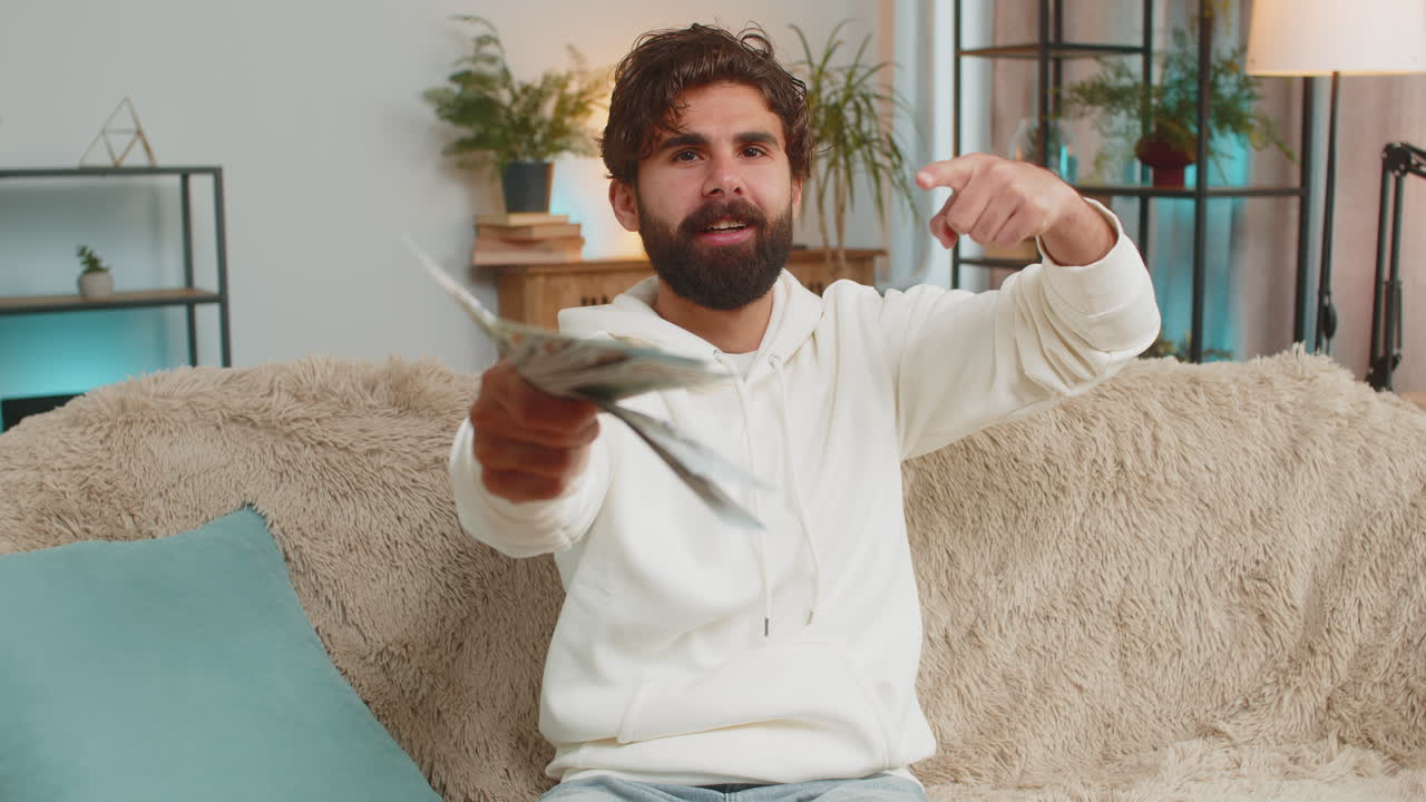 부유한 젊은 히스패닉 남자가 집에서 소파에 앉아 달러 지폐를 들고 있습니다.