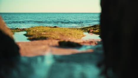 Ocean-shoreline-with-gentle-waves-crash-on-sand-with-seaweed-out-of-focus-between-rocks-at-sunset-4K