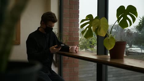 Hombre-Adulto-Joven-Sentado-En-Un-Restaurante-Usando-Un-Teléfono-Inteligente-Con-Una-Máscara-De-Prevención-Covid