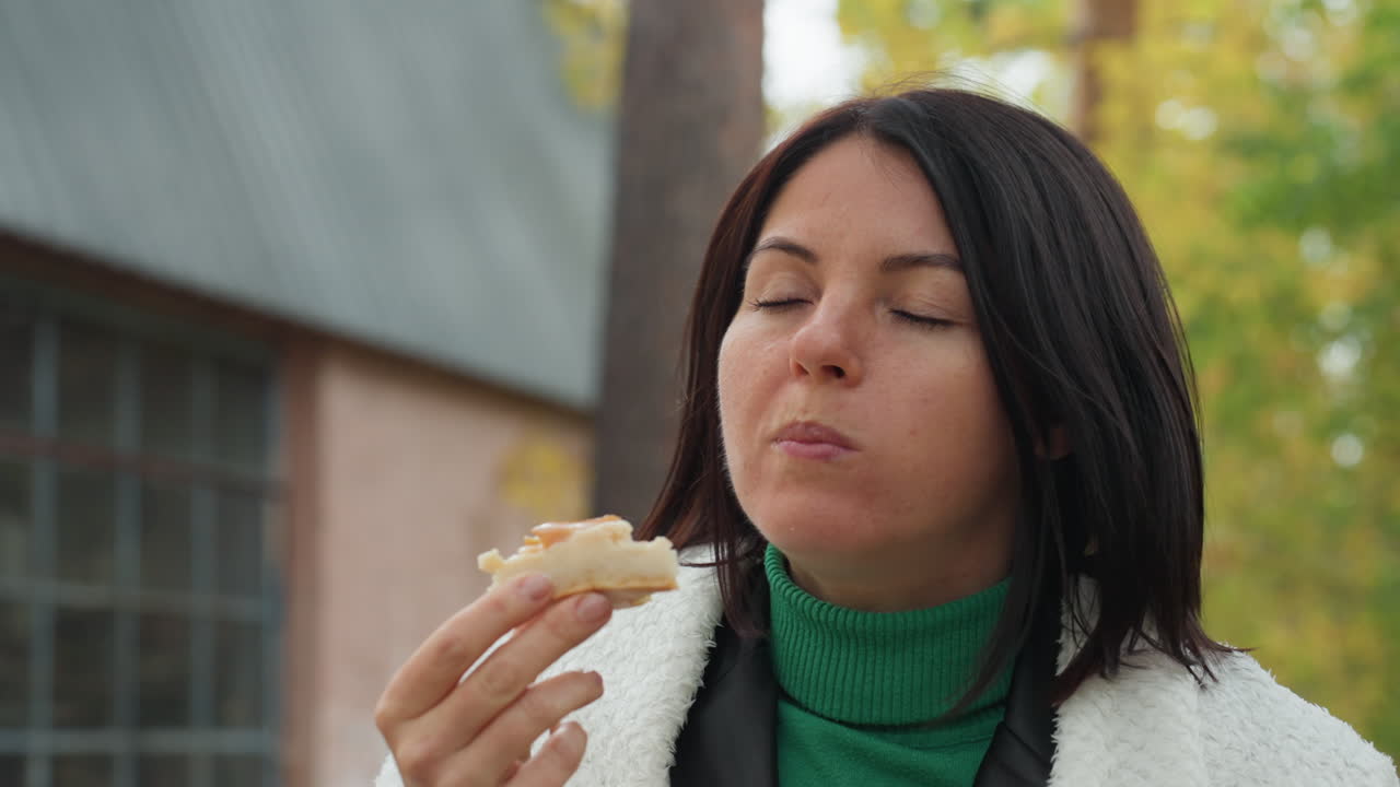 brunette woman eating pastry outdoors in green turtleneck and white fleece coat, inspects pastry, takes bite, sips from metal mug, savors flavor among fall foliage near rustic cottage, candid relaxed