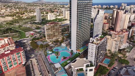 slow drone ascent reveals the skyline of calp, spain, with high-rise apartments, estimar hotel, rooftop pools and a colorful public water park, with the coast and lagoon visible in the background