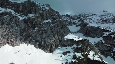 Close-flight-at-a-glacier-rock-snow-mountain-top-near-Bavaria-Elmau-castle-in-the-Bavarian-Austrian-alps-on-a-cloudy-and-sunny-day-along-trees-and-forest-in-nature-with-avalanches-going-down