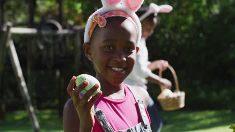 african american girl wearing easter bunny ears doing easter egg hunt in garden with brother