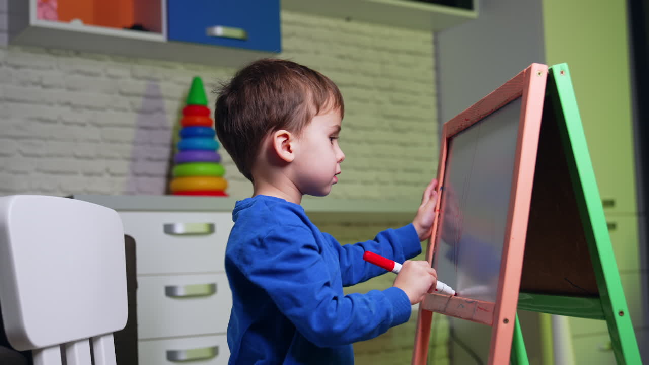 Cute toddler boy wearing blue sweater draws on the blackboard with red marker. Active kid having art hobby at home. Side view.