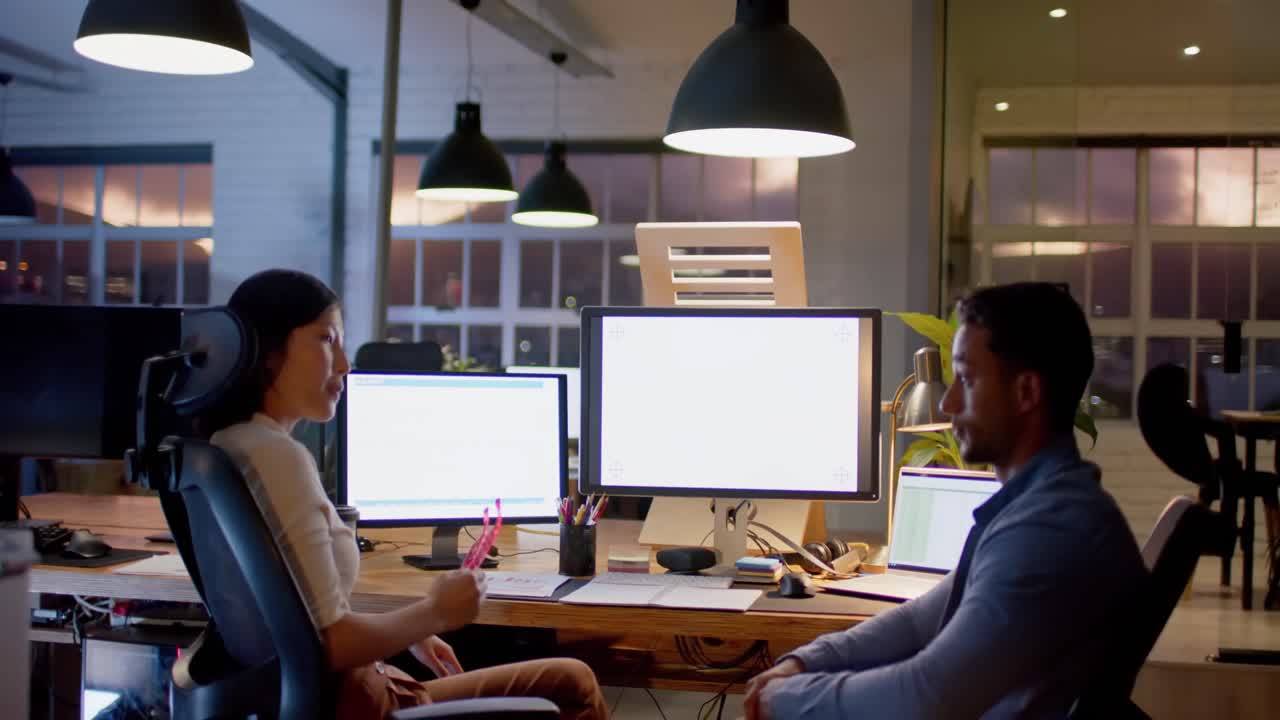 Diverse male and female colleague talking and using computers with copy space at night in office