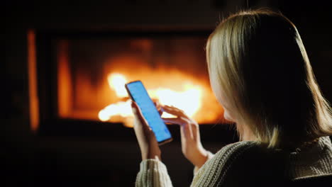 una mujer usa un teléfono inteligente por la noche sentada junto a la chimenea