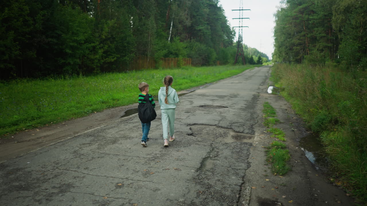 Two children walking side by side on cracked paved road surrounded by green vegetation, engaging in warm conversation with visible potholes, overcast weather, and distant utility poles