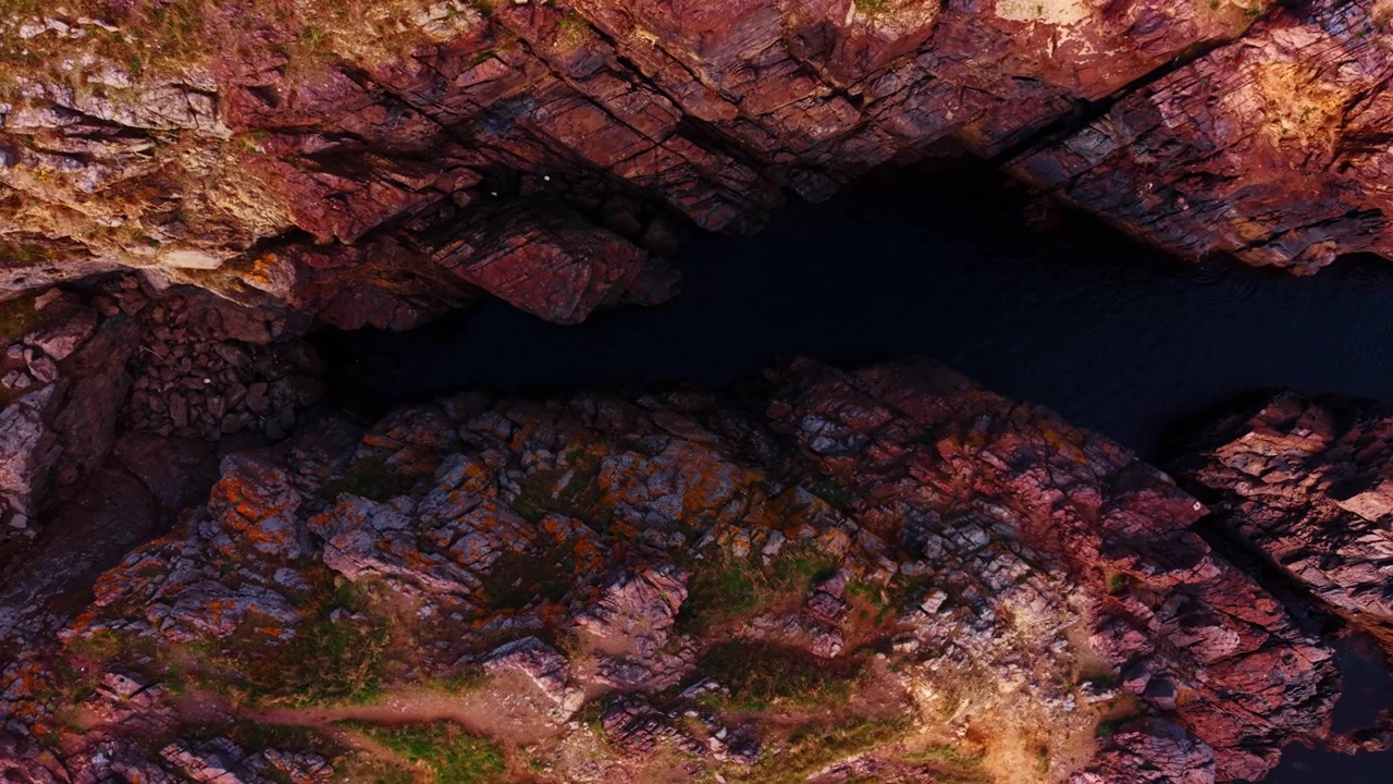 Vista dall'alto di un crepaccio costiero roccioso con una pozza d'acqua scura. Ripresa aerea con drone di roccia stratificata rossa e beige, massi sparsi e bordi erbosi.