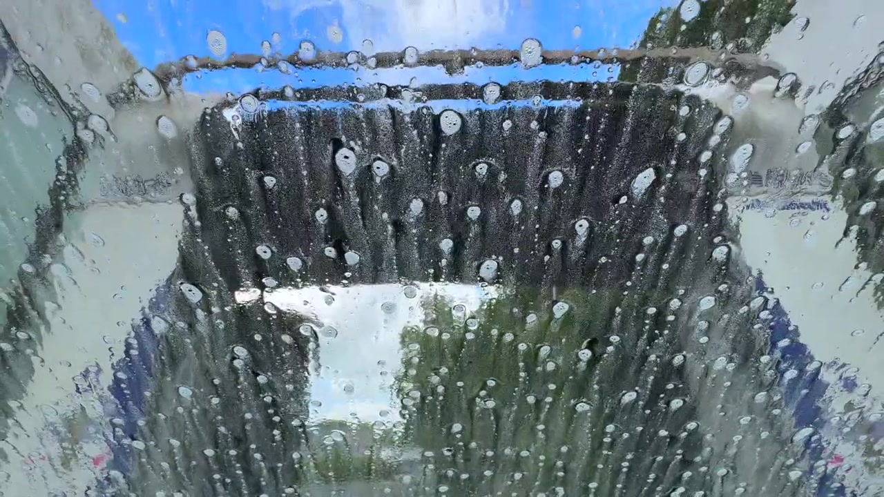 Uma vista através do para-brisa de um carro passando por uma máquina de lavagem automática com escovas giratórias, água e espuma de sabão.