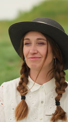 una mujer alegre con el cabello trenzado, con un vestido blanco y un sombrero negro, sonríe mientras está sentada casualmente en un campo verde exuberante