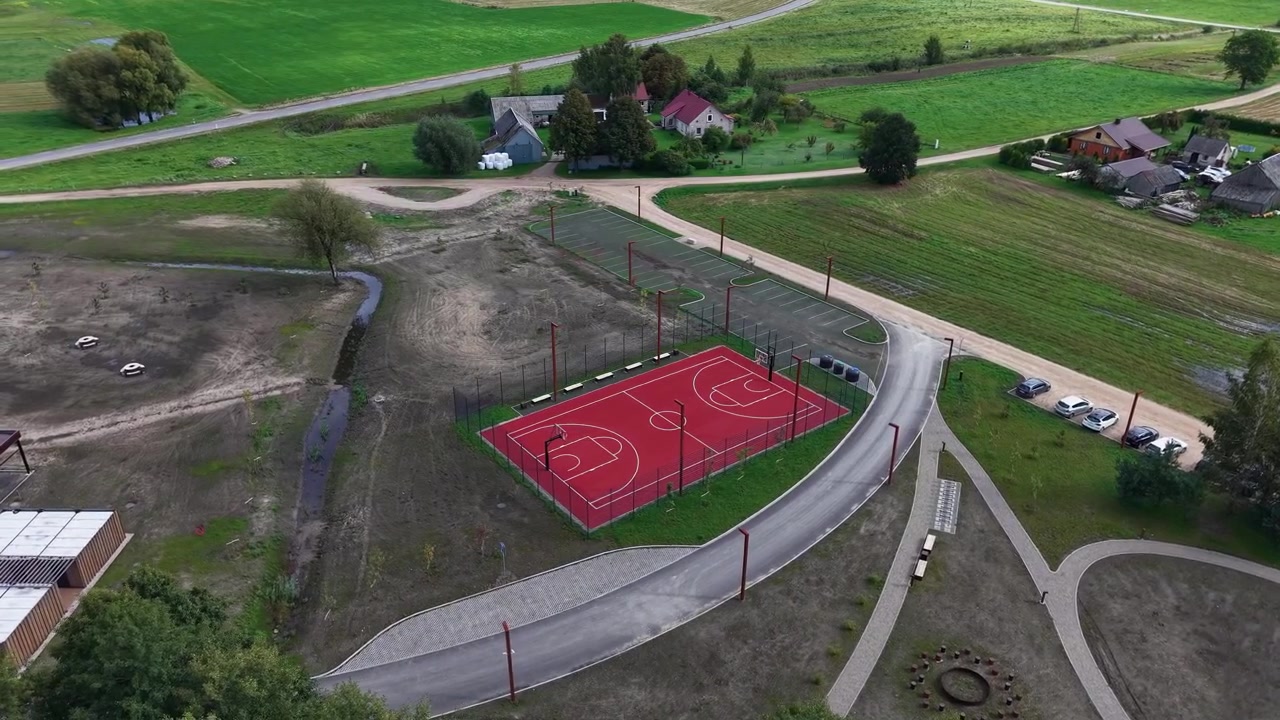 Riprese con drone mostrano un vivace campo da basket rosso immerso in una tranquilla zona rurale, circondato da campi verdi, case sparse e strade tortuose sotto una luce diurna soffusa. Nessuna persona visibile
