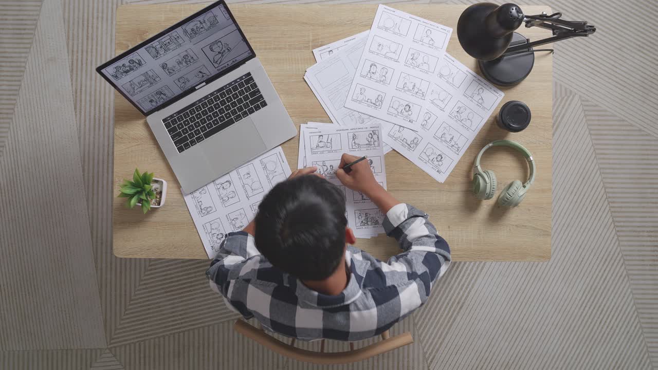 vista superior de un artista masculino asiático con dolor de espalda mientras dibuja un guión gráfico para la película en la mesa con una computadora portátil y auriculares en el estudio