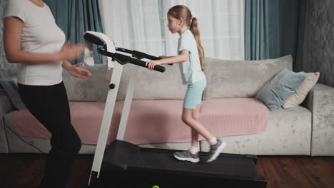 young girl in activewear walks confidently on treadmill while adult offers guidance beside her, home setting features pastel decor, cushioned sofa, and daylight filtering through curtains
