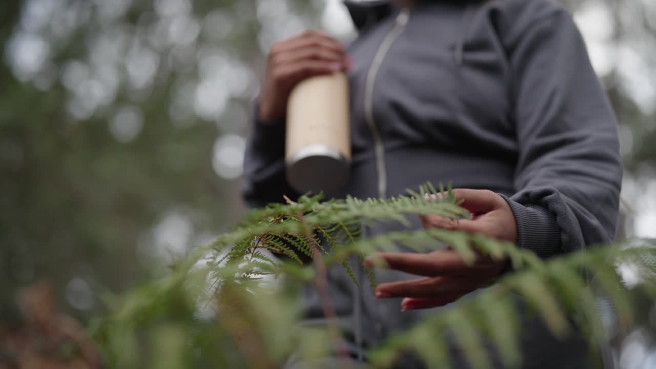 ワークアウトウェアを着て、水筒を持った女性が、シダ植物の緑の葉を優しくなでている。