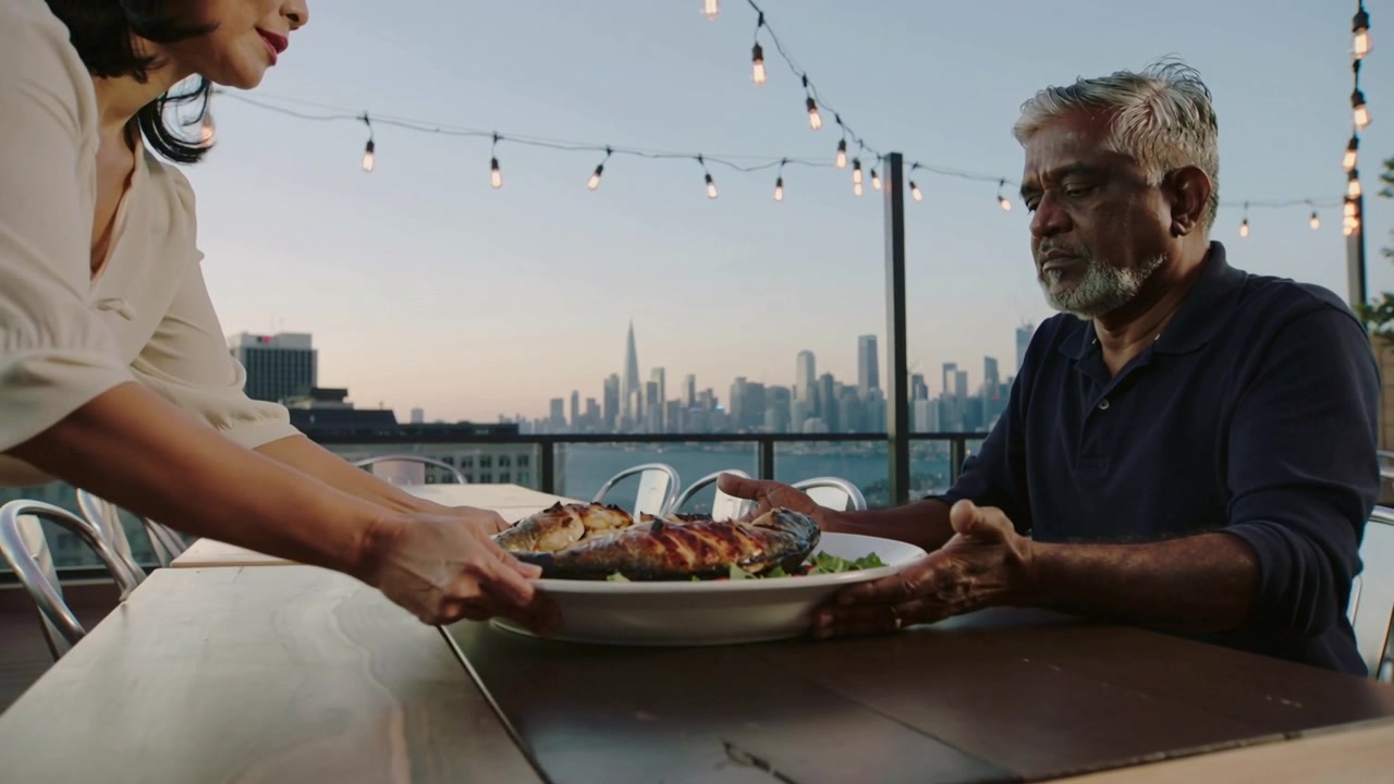 Approaching left Asian woman placing dish onto rooftop table, man reaching to hold dish