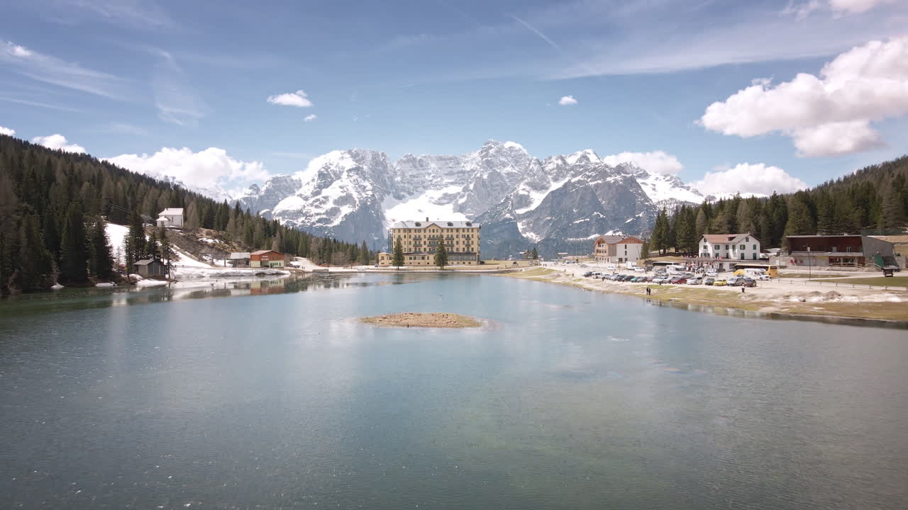 Camera flies straight ahead over a sparkling alpine lake toward a grand lakeside hotel and clustered lodges, framed by dense forest and towering snow-covered peaks