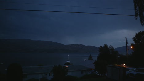 Lightning-storm-flashes-in-clouds-at-night-over-lake-and-mountain-town,-Canada