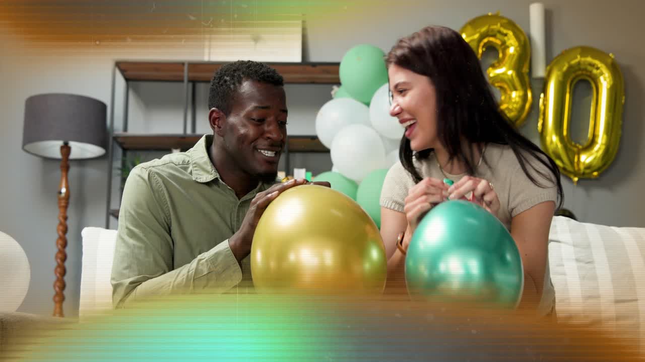 Couple sitting on couch inflating metallic balloons for 30th birthday party, exchanging smiles