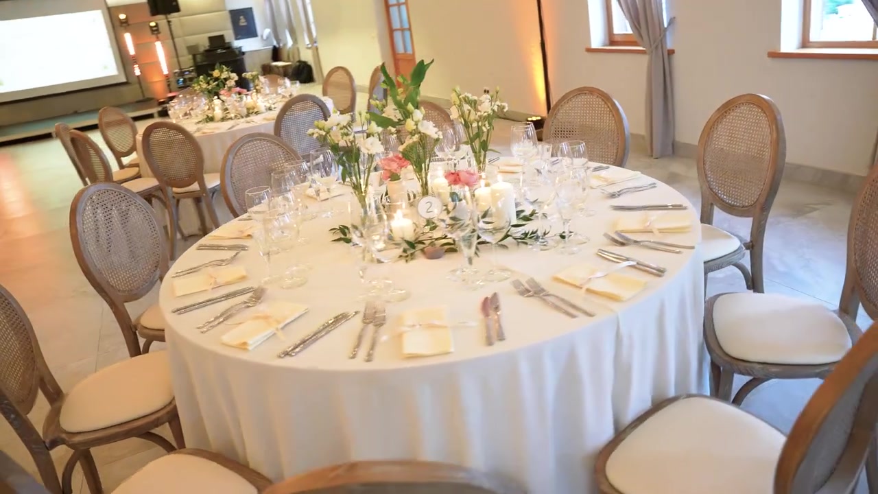 Beautifully arranged round tables with floral centerpieces, candles, fine glassware, and cutlery in a softly lit, empty banquet hall prepared for an elegant wedding or event. (200 characters)