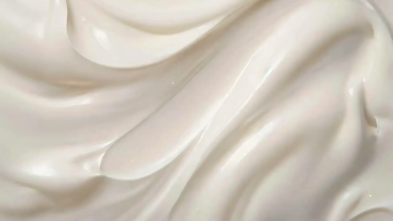 Detailní video záběr hladké, krémové textury s jemným osvětlením, který zobrazuje plynulé, vlnité vzory