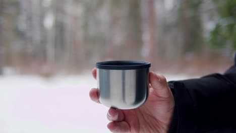 hand holding a metal cup in a snowy forest