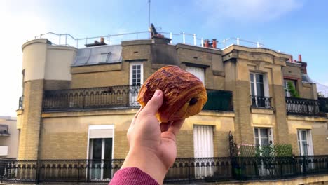 mano sujetando y mostrando pain au chocolat contra edificios parisinos en segundo plano.