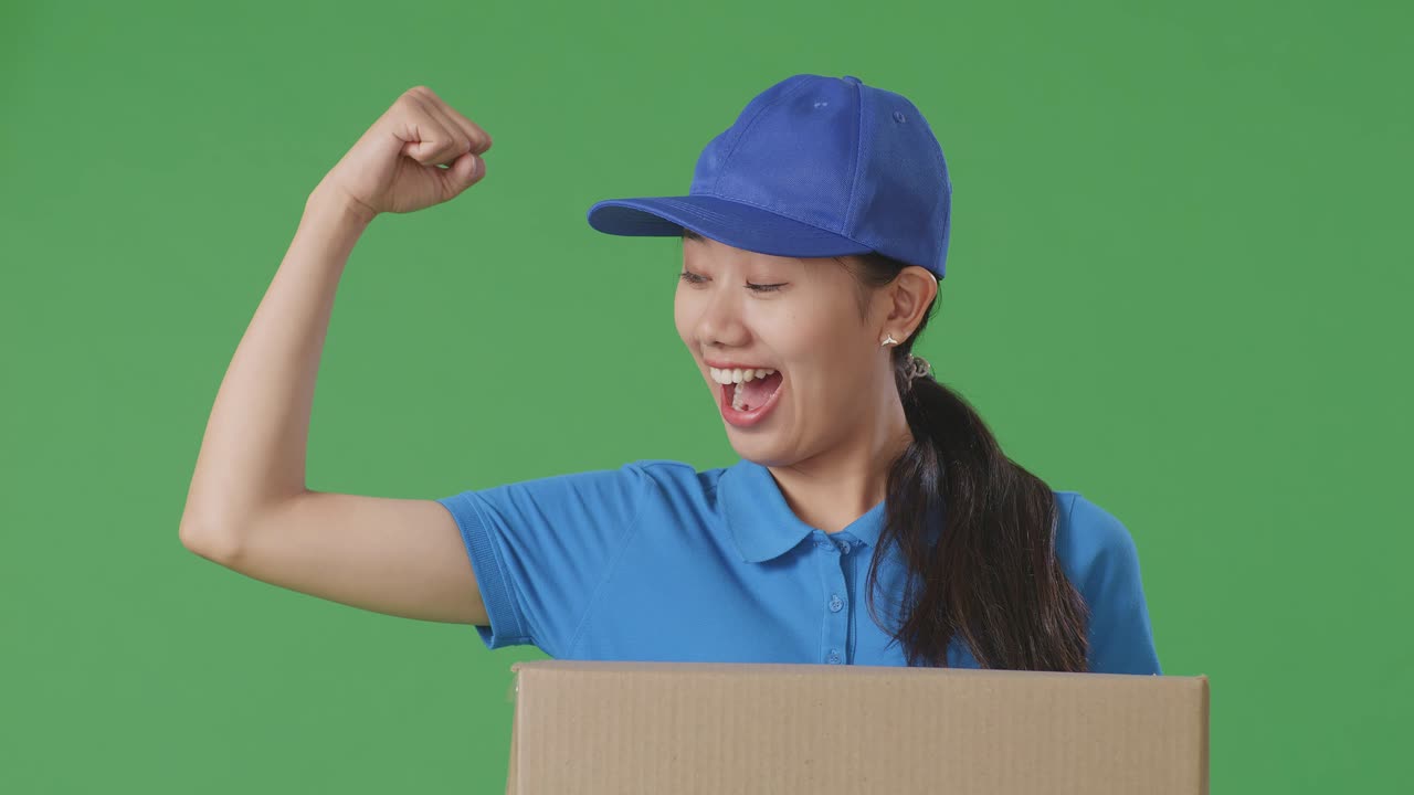 푸른 유니폼을 입은 아시아 여성 택배사의 클로즈업 미소와 플렉스 근육은 스튜디오에서 녹색 화면 배경에 상자를 배달하는 동안