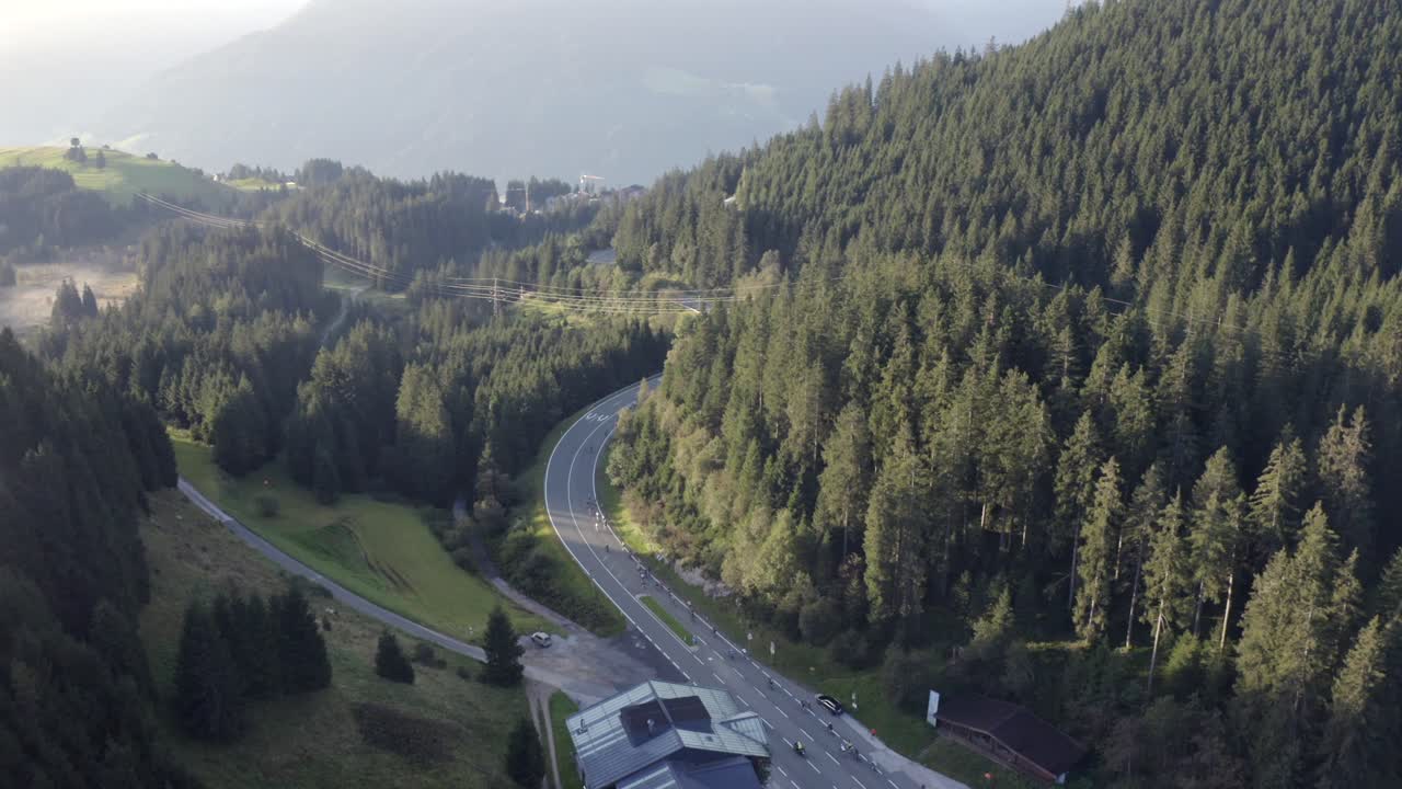 A line of cyclists snakes through a vibrant green alpine meadow beneath traditional Tyrolean homes.