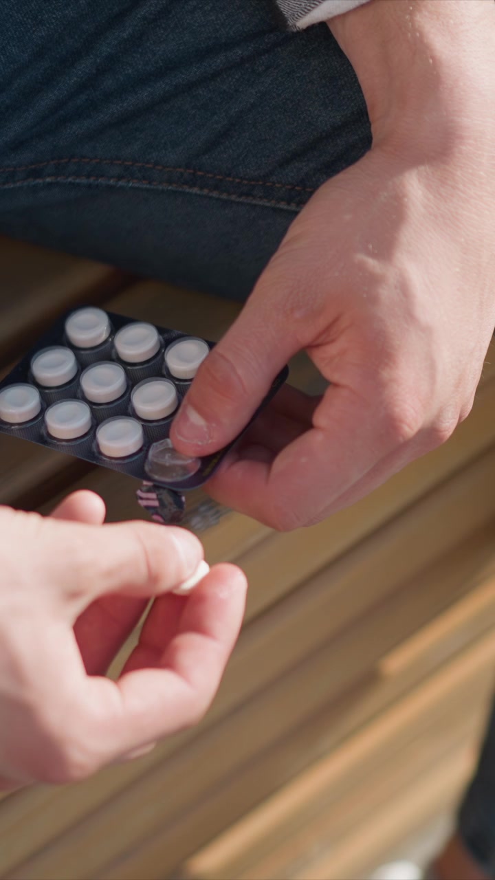 Close up of hands holding blister pack of white round tablets one person tearing open foil seal while another holds package. Hands are shown in detail against background with wooden slats