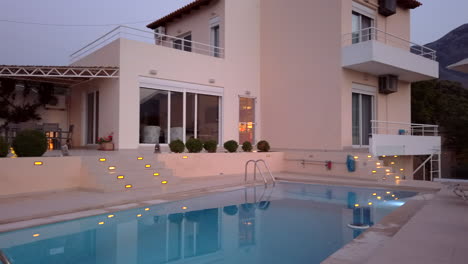 exterior pan shot of luxury greek villa at dusk with outside lighting and pool in foreground with loungers