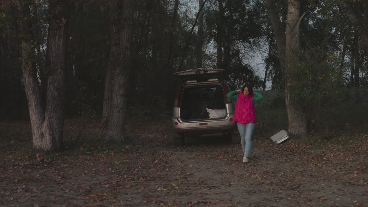 Person unloading car at dusk, open trunk by trees and cardboard box on ground, pink jacket and jeans, setting up small campsite beside forest clearing, quiet twilight atmosphere with fallen leaves