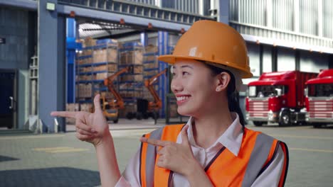 Close Up Of Asian Female Engineer With Safety Helmet Smiling And Pointing To Side, Outside of Logistics Distributions Warehouse
