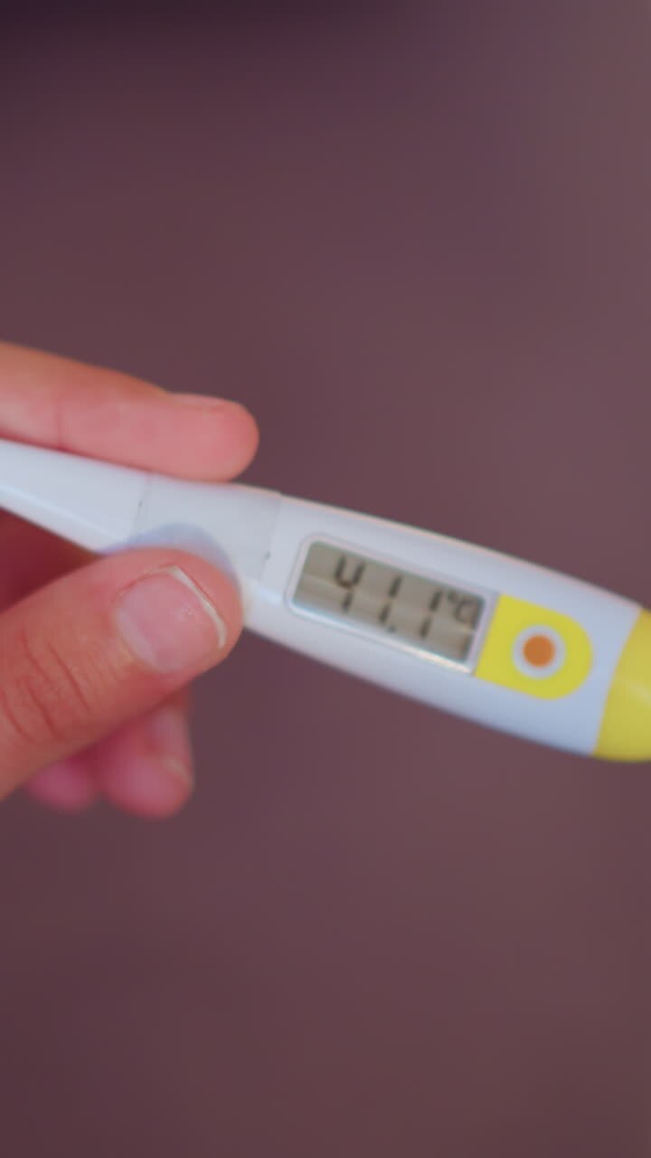 Nærbillede af hænder, der holder et digitalt termometer, som viser 41,1 grader Celsius, mens den anden hånd delvist dækker displayet, hvilket antyder bekymring, hast og opmærksomhed omkring høj feber