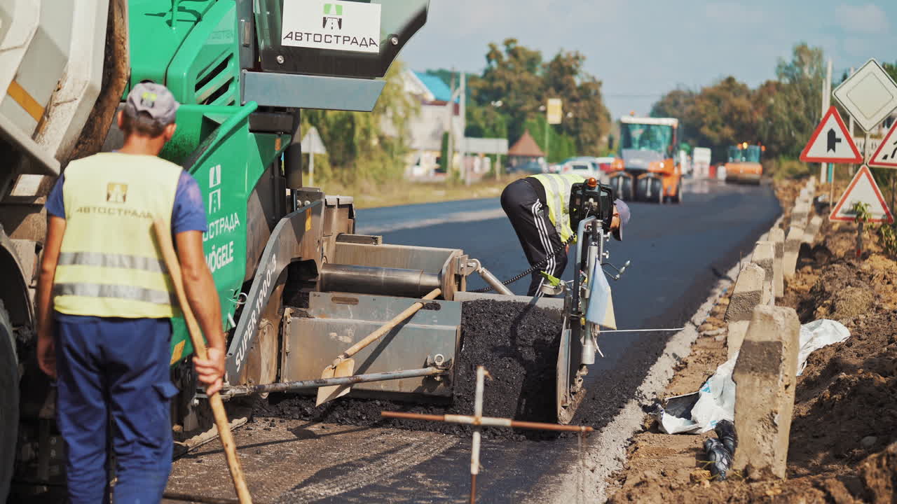Asphalt machine lays asphalt on highway on the background of heavy machinery at day. Workers in protective uniform control the work of asphalt paver machine during road construction.