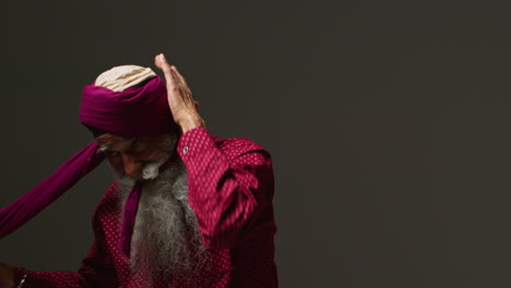 low key studio lighting shot of senior sikh man with beard tying fabric for turban against dark background