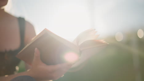 woman seated outdoors holding book, flipping through pages as sunlight creates a glowing ambiance around it, background is softly blurred