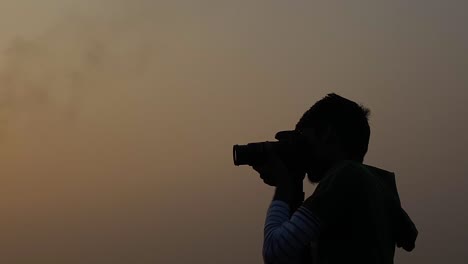 Silueta-De-Un-Joven-Fotografiando-Con-Una-Cámara-Dslr-Contra-El-Cielo-Naranja