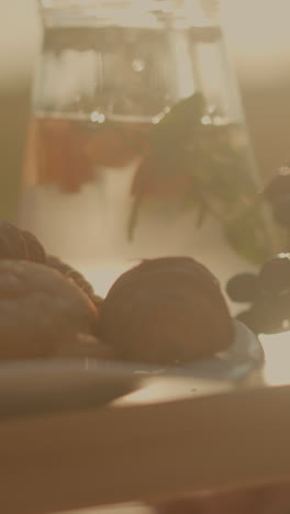 primer plano de la configuración de picnic rústico en la mesa de madera con croissants frescos, uvas verdes y frutas variadas como rebanadas de naranja y melocotón, junto a la jarra de agua bajo dorado
