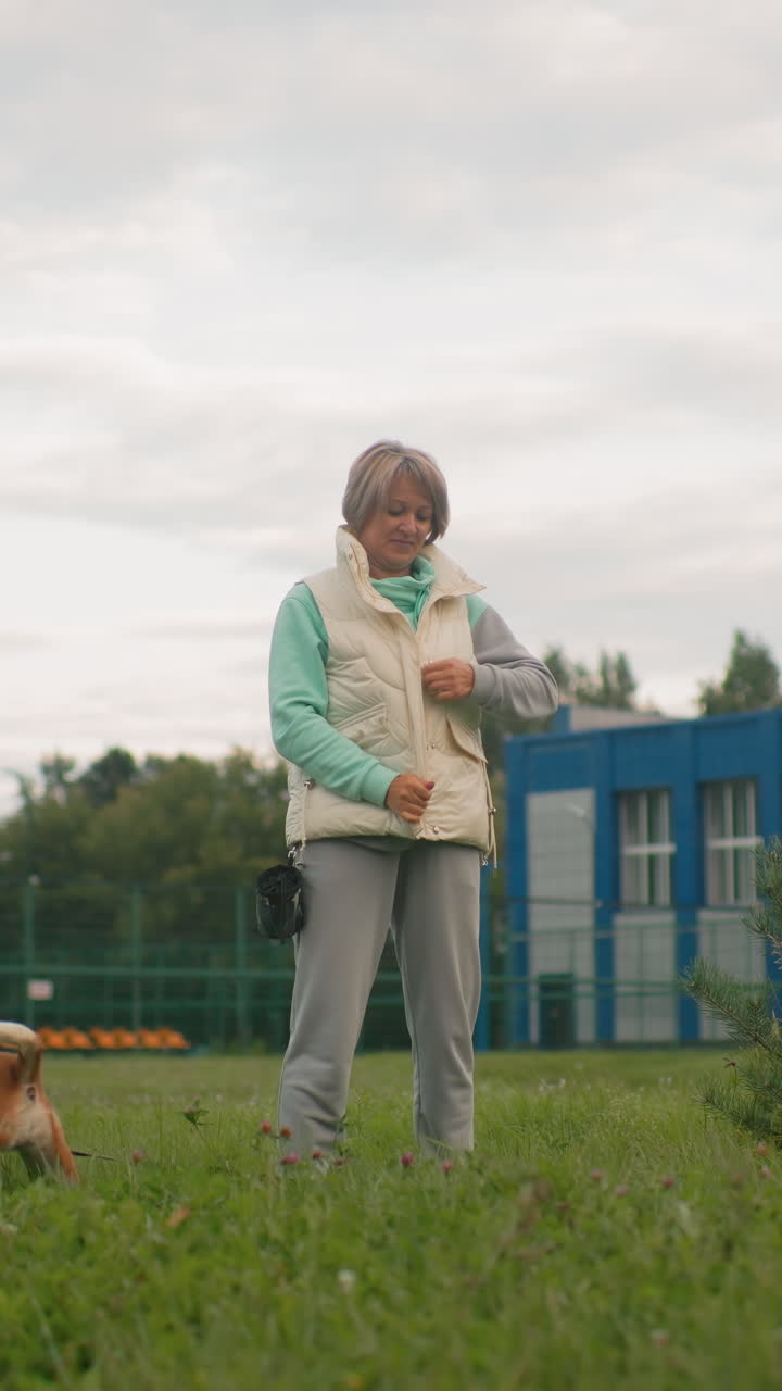 Older white woman standing in park, retiree enjoying quiet stroll with beagle at feet, wearing puffy vest and hoodie, grassy field and blue sports building in background, overcast sky, calm reflective