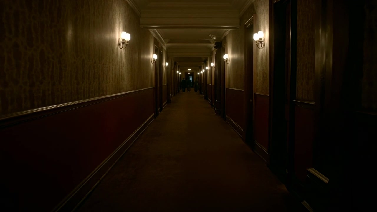 Corridoio d'hotel in penombra, profonda prospettiva, pareti con carta da parati vintage, applique a parete che proiettano pozze di luce calda.