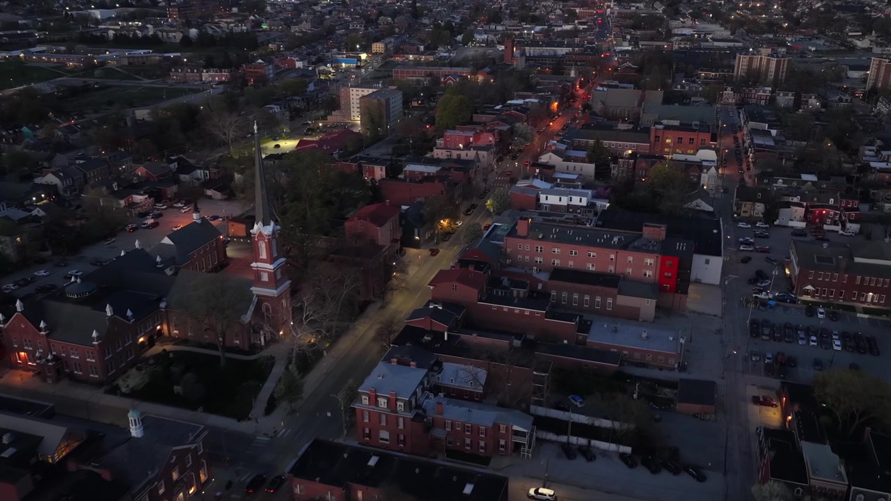 Charming American city at dusk with glowing streets, historic brick buildings and church steeple. Aerial flyover reveals peaceful downtown blocks, rooftops and warm evening lights. Slow drone top view