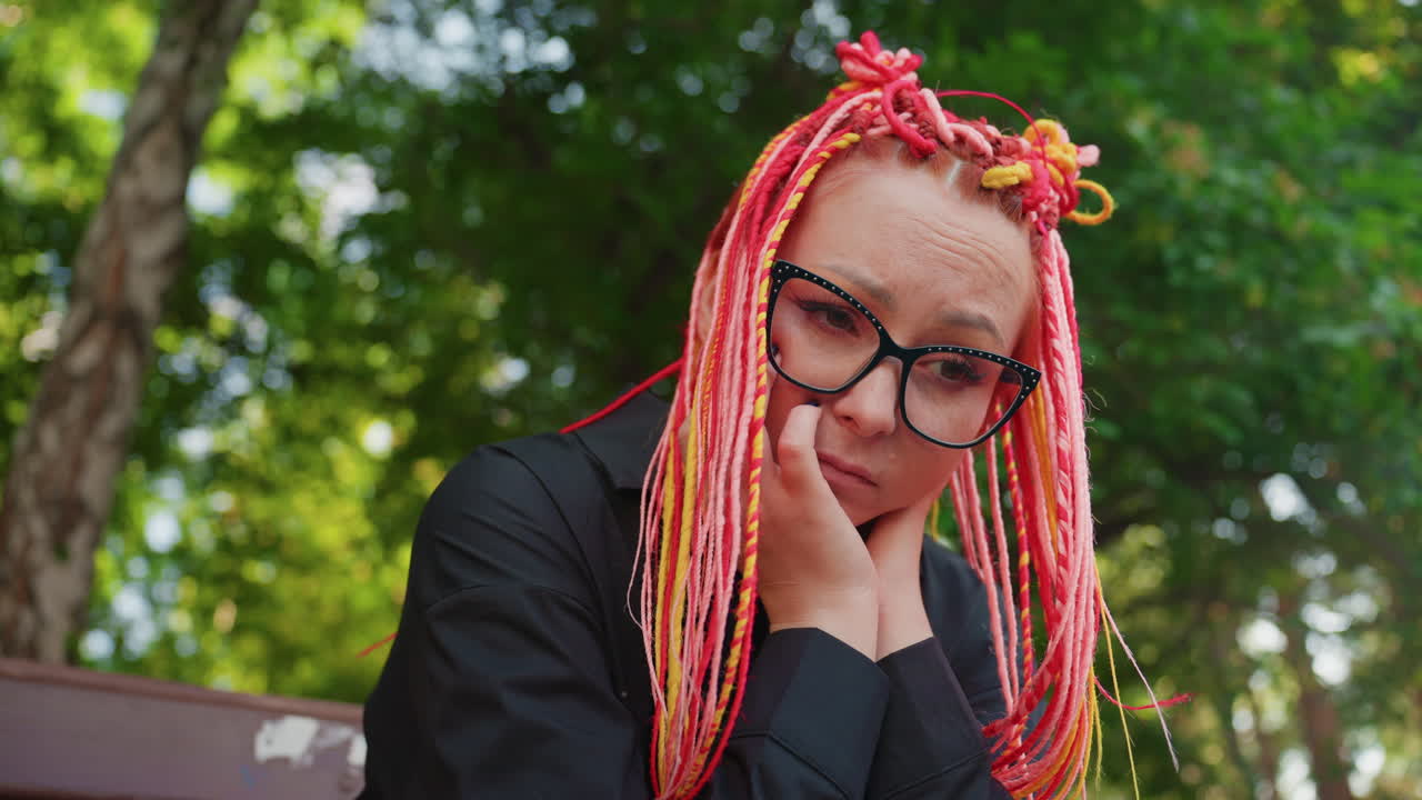 공원 벤치에 앉아 생각에 잠긴 백인 여성, 핑크색과 노란색 땋은 머리, 커다란 안경, 턱을 손에 괸 모습, 부드러운 햇빛과 푸른 나무, 캐주얼한 검은색 재킷, 창의적인 학생 분위기, 내성적인