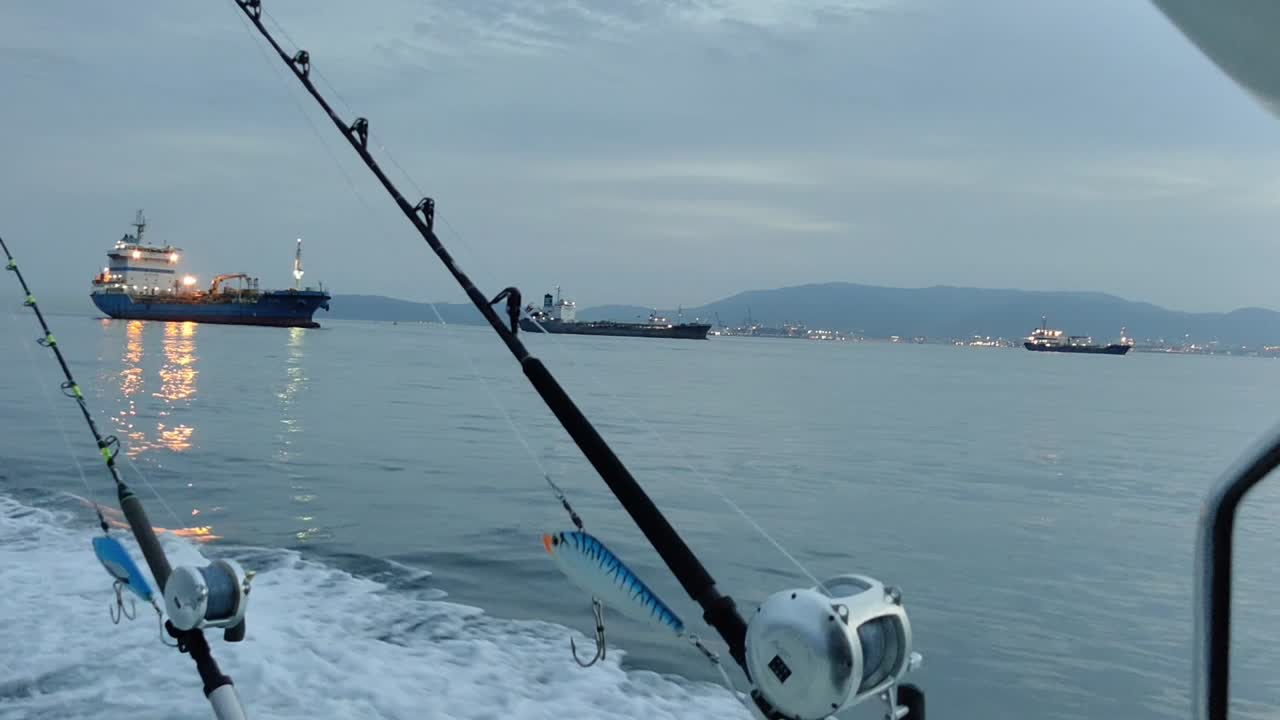 View of the Fishing boat with Two fishing rods mounted on rod holders, swimming in the waters of the Strait of Gibraltar and the cargo or cruise ships in the background
