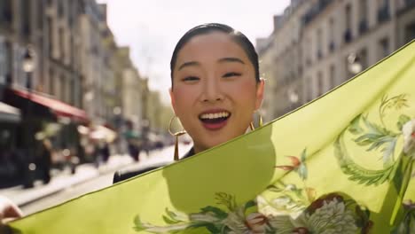 a woman smiles brightly while holding a colorful scarf in the charming streets of paris. the lively atmosphere features sunlit cafes and bustling pedestrians, creating a joyful ambiance.
