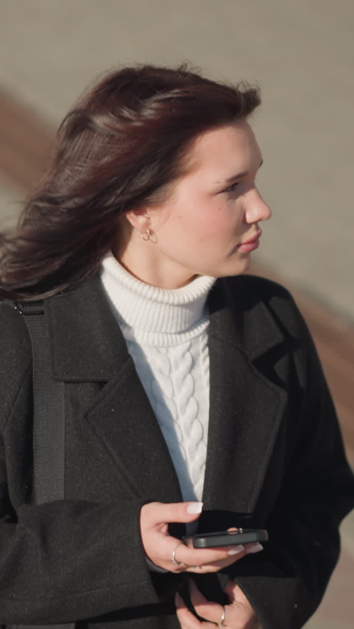 혀있는 길에서 야외를 고 있는 여인, 머리카락을 만지고, 손에 드폰을 들고, 도시 풍경을 돌아다니면서 주변을 둘러보고