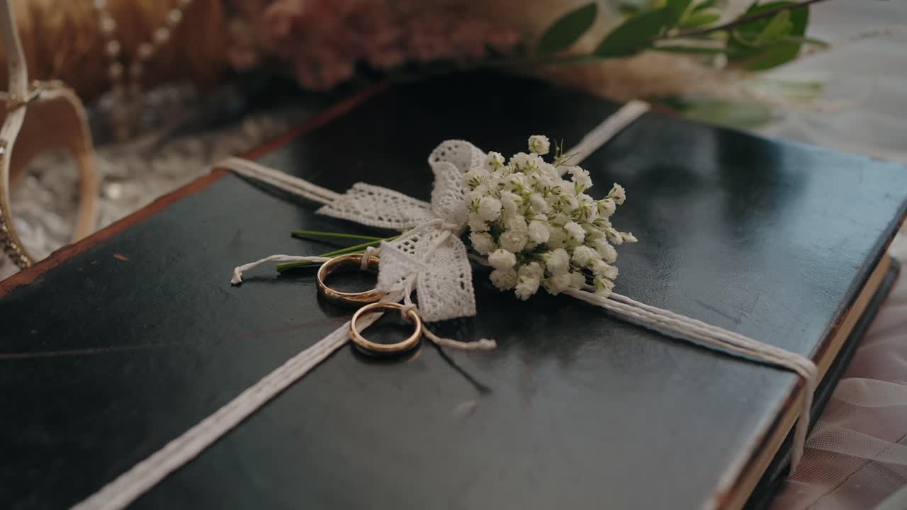 Two gold rings on a black Bible tied with lace and white flowers in soft natural light