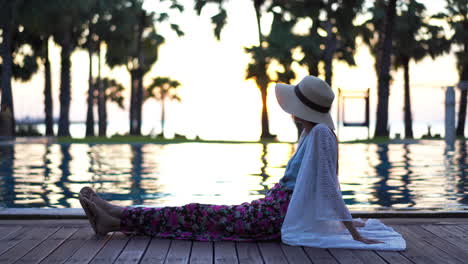 a young woman casually dressed sits on the wooden decking surrounding a resort swimming pool looking out to the ocean