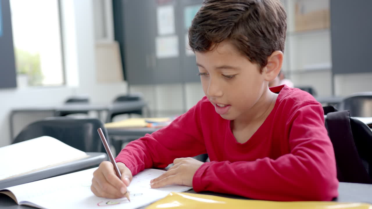 In school, boy wearing a red shirt writing in a notebook in a classroom
